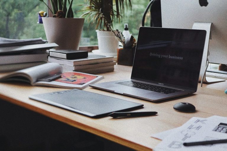 Online - Homeoffice Arbeitsplatz mit Laptop, Tablet, Stiften und Zeitschriften auf einem Holztisch.
