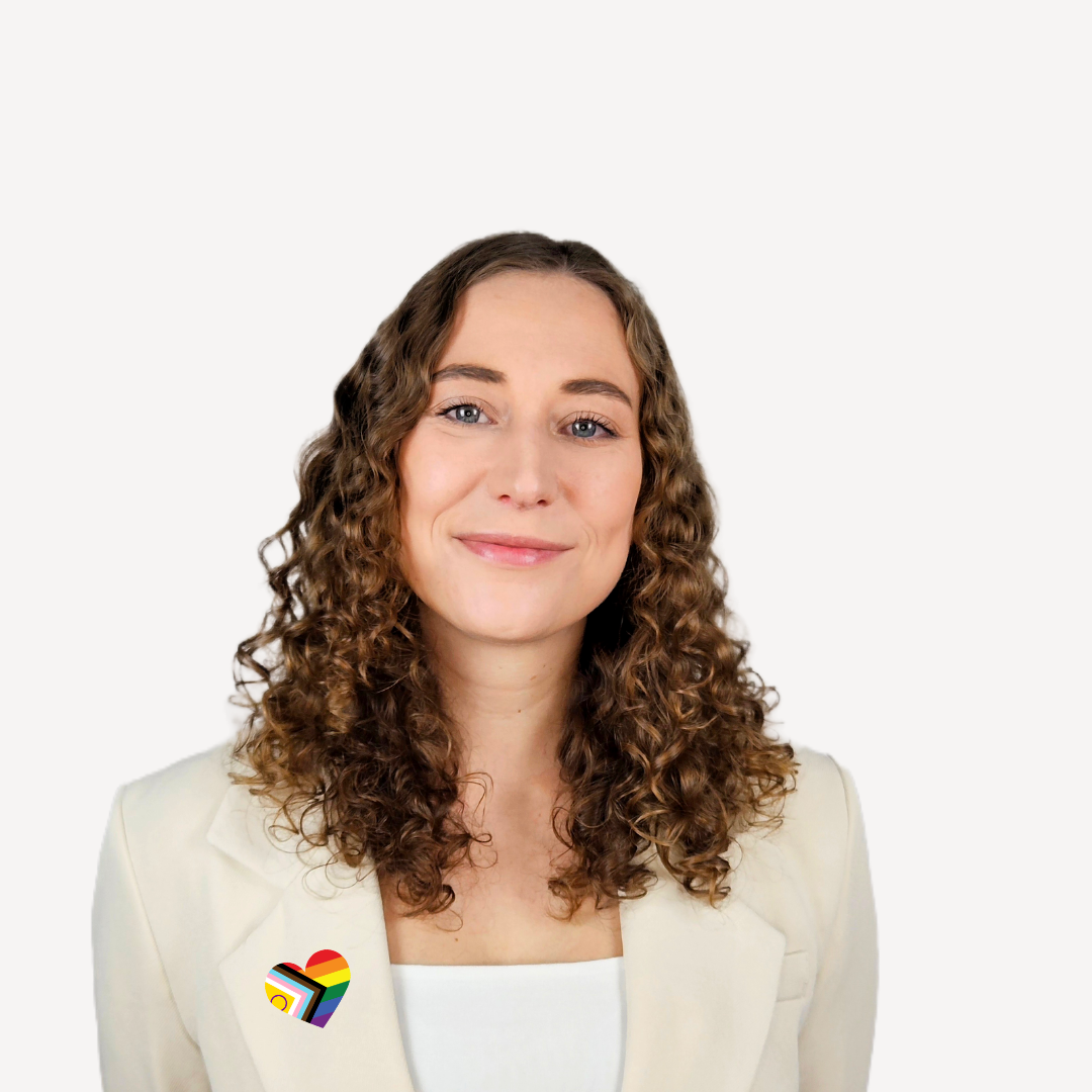 Portrait Hannah Sophie Welte Lächelnde Person mit lockigem Haar, trägt ein helles Outfit und ein Regenbogen-Emblem der Progress Pride Flag in Form eines Herzens.